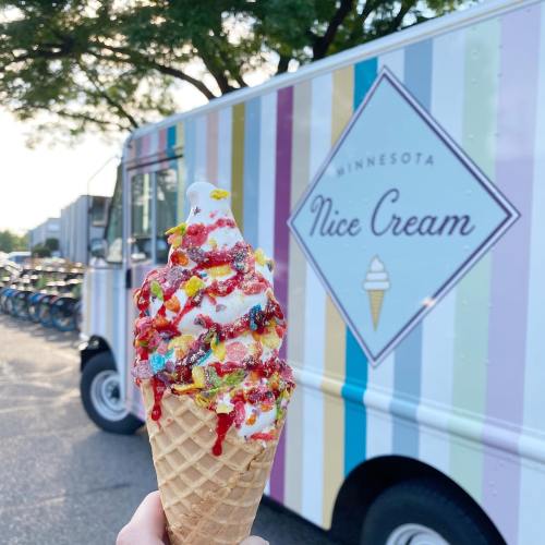 A hand holds a colorful ice cream cone topped with sprinkles and syrup, with a striped ice cream truck in the background.