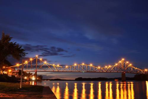 A lit bridge spans a calm river at dusk, reflecting warm lights on the water's surface under a cloudy sky.