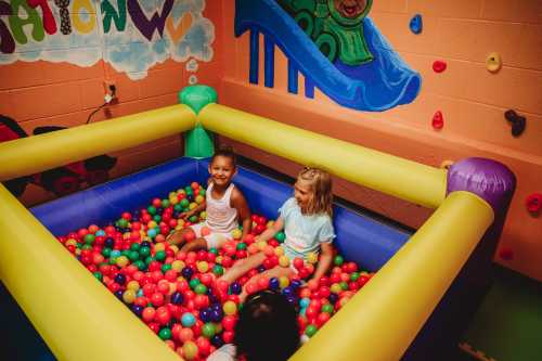 Two children sit in a colorful ball pit, surrounded by vibrant walls and playful decor.