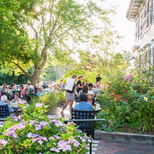 Outdoor dining scene with people at tables surrounded by flowers and trees, enjoying a sunny day.