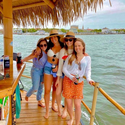 Four women in summer outfits enjoy a sunny day on a boat, smiling and holding drinks by the water.
