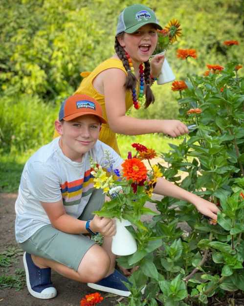 Two children picking flowers in a garden, one holding a vase and the other smiling with a bouquet in hand.