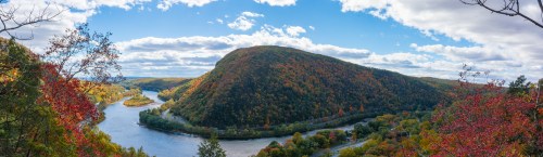 A panoramic view of a river winding through colorful autumn foliage and rolling hills under a blue sky with clouds.