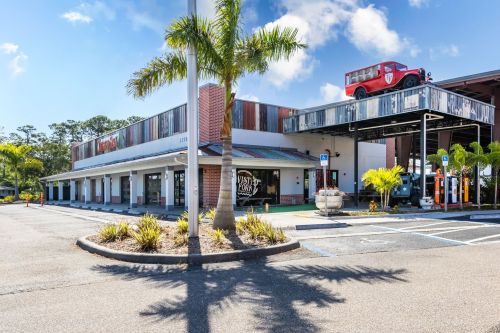 A building with a red truck on the roof, surrounded by palm trees and a parking lot under a clear blue sky.
