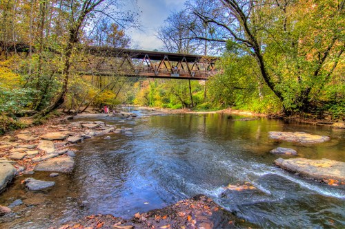 A serene river scene with a bridge overhead, surrounded by lush trees and colorful autumn foliage.