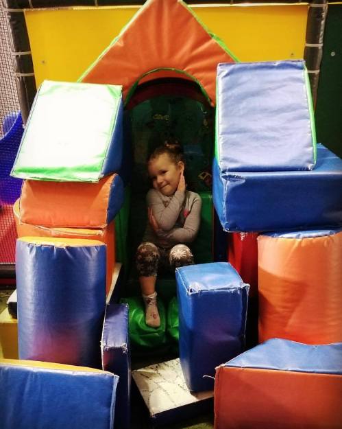 A child sits playfully among colorful foam blocks in a play area, smiling and resting her chin on her hand.