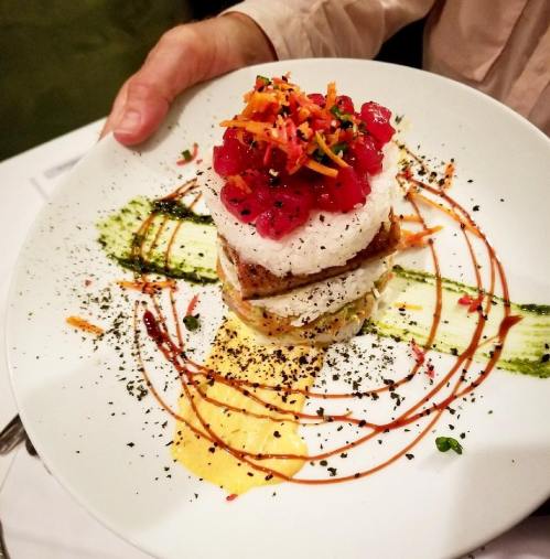 A plate of layered sushi with rice, tuna, and colorful garnishes, drizzled with sauces and herbs.