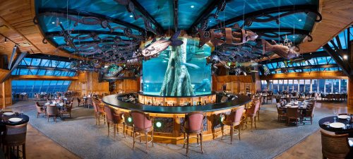 A spacious restaurant with a large aquarium centerpiece, wooden decor, and panoramic windows showcasing a scenic view.