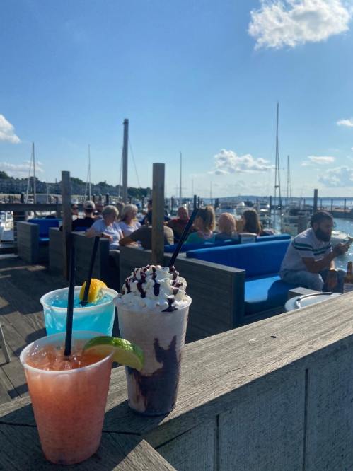 Three colorful drinks on a wooden table with a marina view, people enjoying the outdoors in the background.