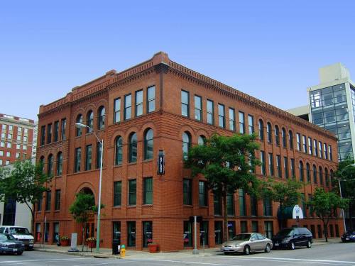 Historic red brick building with arched windows, surrounded by trees and modern buildings in an urban setting.