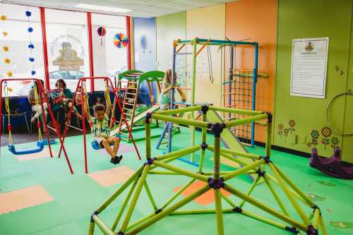 A colorful indoor play area with swings, climbing structures, and children playing on soft flooring.