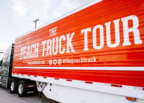 A bright orange truck with "The Peach Truck Tour" written on the side, parked in a lot under a cloudy sky.