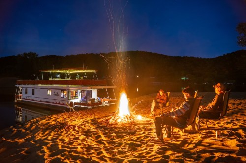 Three people sit by a campfire on the beach at night, with a houseboat illuminated in the background.