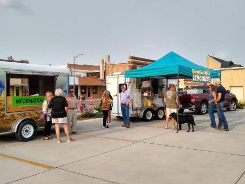 A food truck and lemonade stand with people chatting and a dog in a parking lot on a sunny day.
