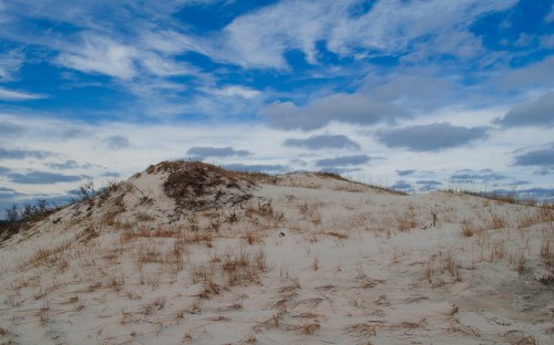 A sandy dune with sparse grass under a blue sky filled with clouds.