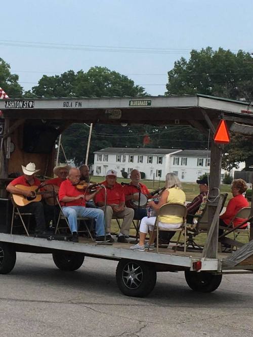 A group of musicians in red shirts playing instruments on a flatbed trailer, with an audience seated nearby.