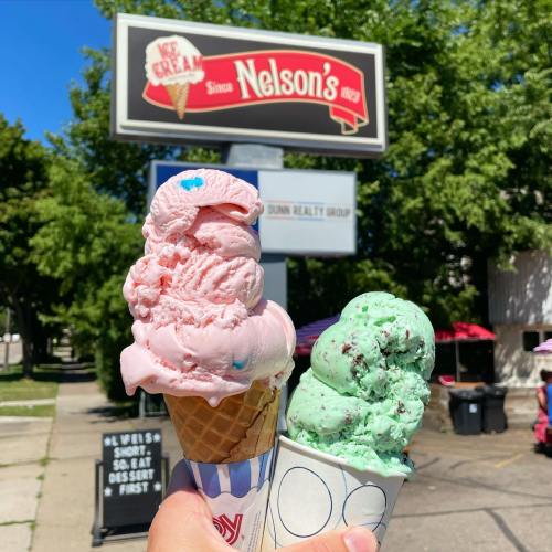 A hand holds two ice cream cones: one with pink ice cream and the other with green mint chocolate chip, in front of a sign.