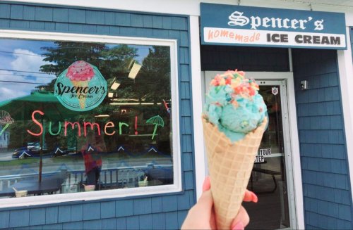 A hand holds a colorful ice cream cone in front of Spencer's Homemade Ice Cream shop with a summer-themed sign.