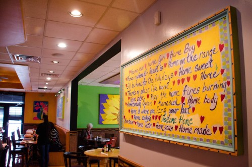 Colorful wall art in a restaurant features heartfelt phrases about home, love, and comfort, with a cozy dining atmosphere.
