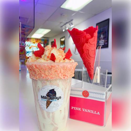 A colorful milkshake topped with whipped cream, sprinkles, and a red cone, displayed in a bright ice cream shop.