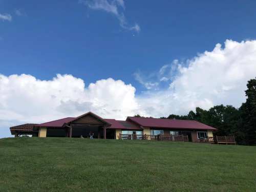 A large house with a red roof sits on a grassy hill under a blue sky with fluffy white clouds.