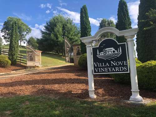 Sign for Villa Nove Vineyards at the entrance, surrounded by greenery and trees under a blue sky.
