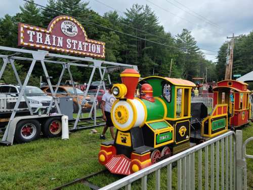 A colorful miniature train at a train station, surrounded by greenery and a sign reading "Train Station."