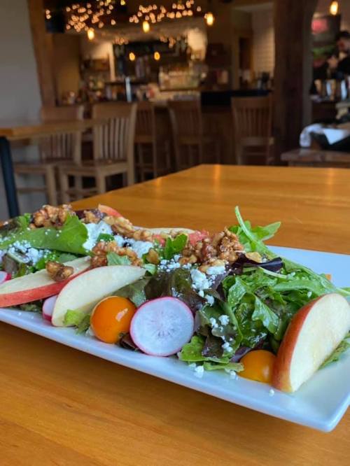 A fresh salad with greens, apple slices, radishes, and walnuts on a white plate, set on a wooden table in a restaurant.
