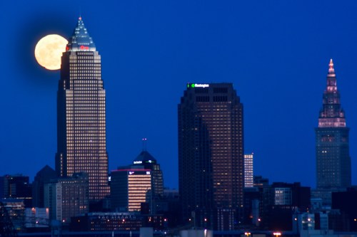 A full moon rises behind Cleveland's skyline, illuminating the tall buildings against a twilight sky.
