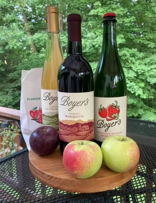 Three bottles of Boyer's wine on a wooden platter, with two apples and a plum, set against a green forest backdrop.