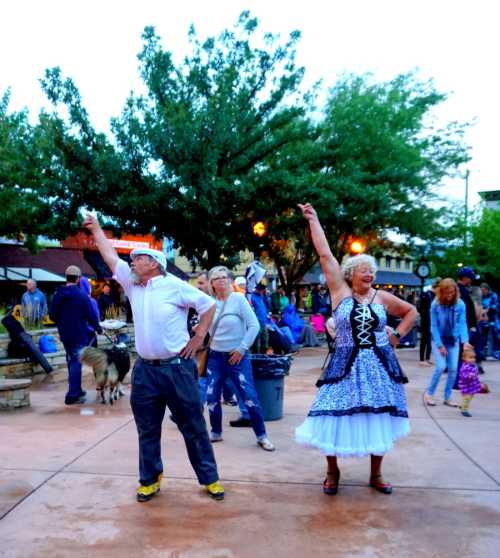 A lively outdoor scene with people dancing joyfully, raising their arms, surrounded by trees and a festive atmosphere.