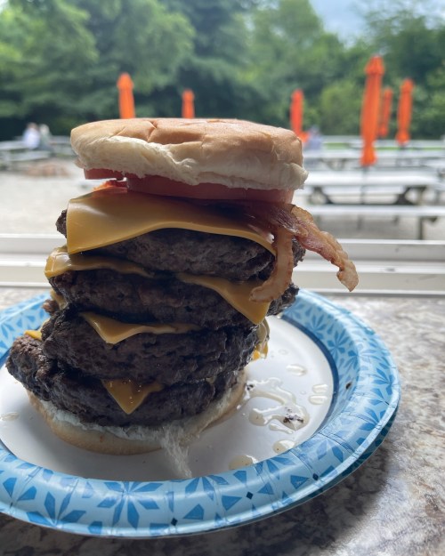A towering burger with multiple beef patties, melted cheese, bacon, and a top bun, served on a paper plate.