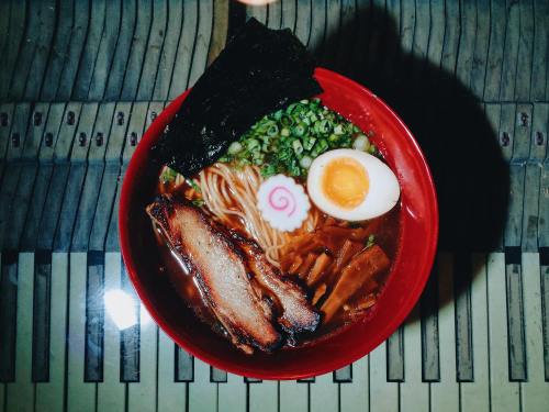 A bowl of ramen with sliced pork, a soft-boiled egg, green onions, and a pink swirl fish cake on a piano keyboard background.