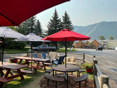 Outdoor seating area with red umbrellas, picnic tables, and a view of mountains in the background.