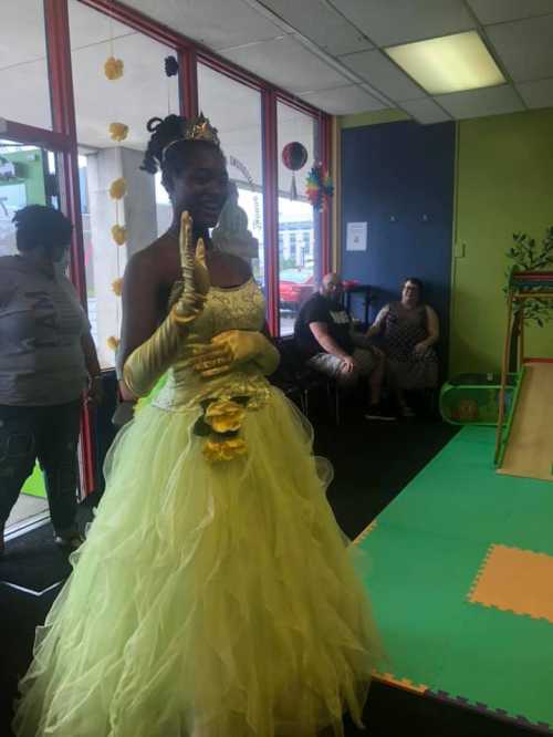 A person in a yellow gown and crown smiles, waving in a colorful room with seated guests in the background.