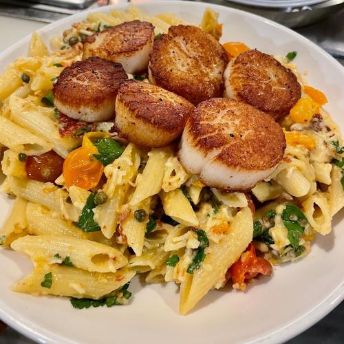 A plate of penne pasta topped with seared scallops, cherry tomatoes, and fresh herbs in a creamy sauce.