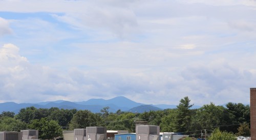 A scenic view of mountains under a cloudy sky, with trees and buildings in the foreground.