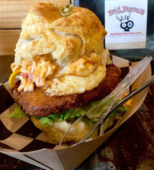 A towering biscuit sandwich filled with fried chicken, egg, and toppings, served in a paper tray.