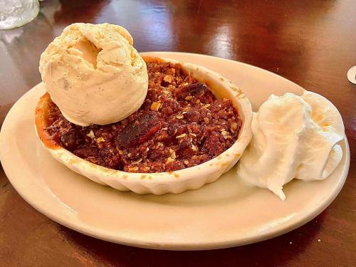 A dessert dish featuring warm cobbler topped with a scoop of vanilla ice cream and a dollop of whipped cream.