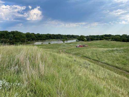 A grassy hillside overlooks a river, with trees lining the bank and a red vehicle parked nearby under a cloudy sky.
