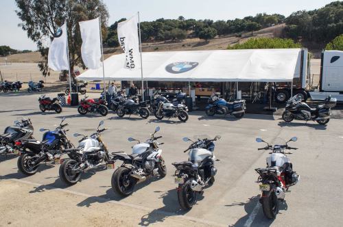 A lineup of BMW motorcycles in front of a large tent at an outdoor event, with trees and hills in the background.