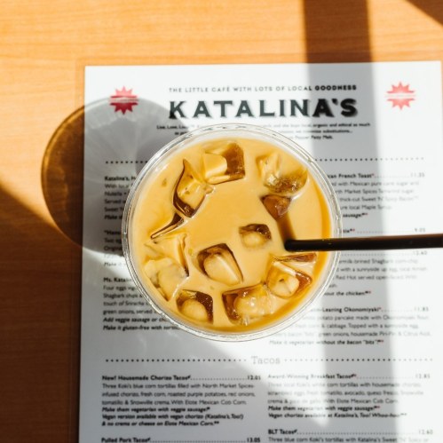 A glass of iced coffee with ice cubes sits on a menu at Katalina's café, illuminated by sunlight.