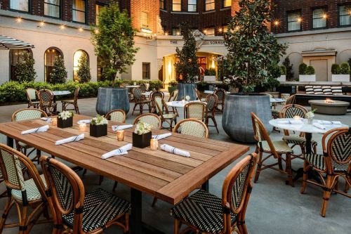 Outdoor dining area with wooden tables, chairs, and greenery, set for guests at a stylish restaurant.