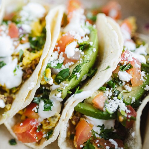 Three tacos filled with meat, avocado, tomatoes, cilantro, and drizzled with sour cream and cheese.