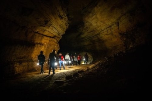 A group of people walking through a dark cave, holding flashlights, with rocky walls illuminated by their lights.