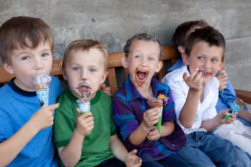 Four children sitting on a bench, enjoying ice cream cones and making playful faces with chocolate smeared on their mouths.
