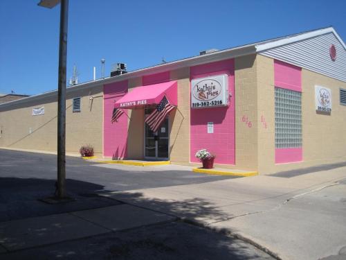 A pink storefront with "Kathy's Pies" sign, featuring American flags and flower pots outside on a sunny day.