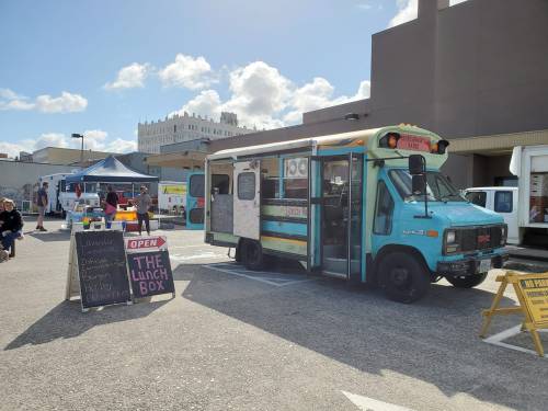 A colorful food truck named "The Lunch Box" parked at a market, with a sign indicating it's open for business.