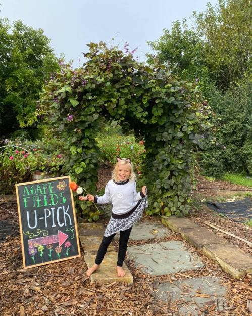 A child in a playful outfit stands by a sign for a U-Pick farm, surrounded by greenery and flowers.
