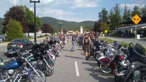 A busy street lined with various motorcycles and people, set against a backdrop of trees and mountains.